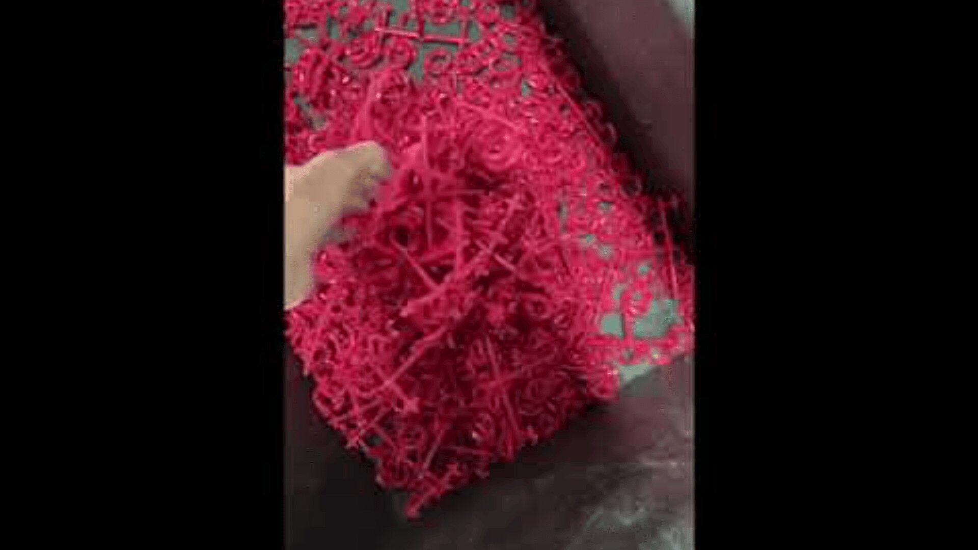 Eine Hand hält einen wirren Haufen kleiner, roter Plastikteile in verschiedenen Formen, möglicherweise Teile aus einem Herstellungsprozess, über einen Behälter, der mit weiteren gleichen Gegenständen gefüllt ist.