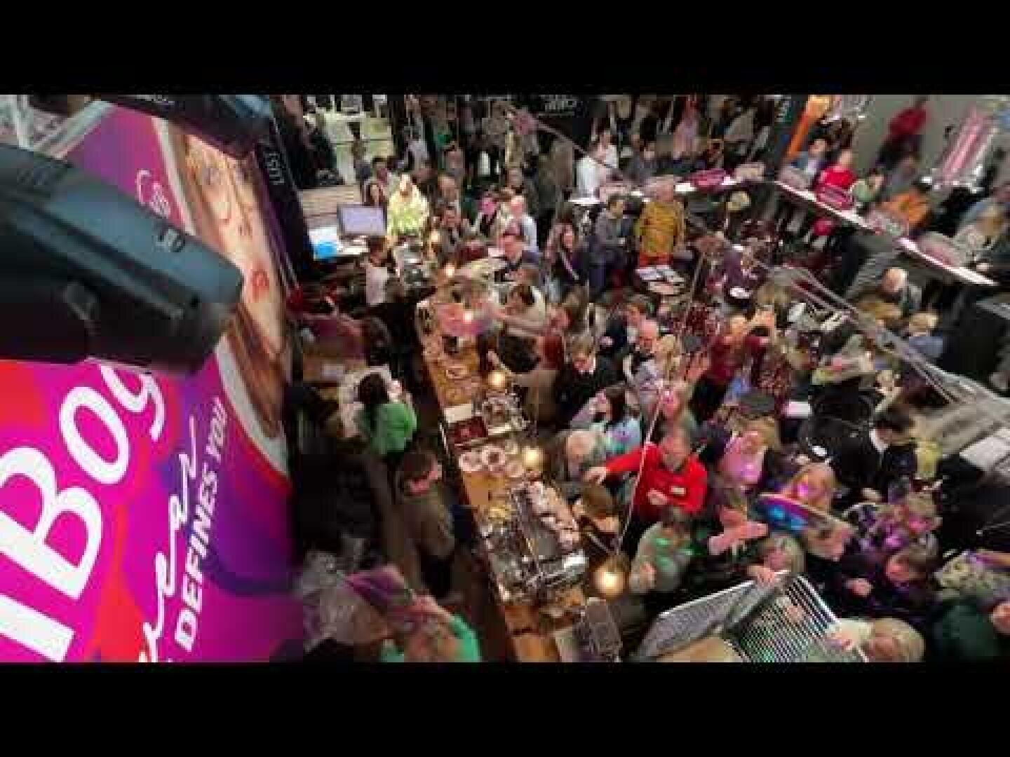 Eine überfüllte Markthalle mit vielen Menschen, die sich um Essensstände scharen, von oben betrachtet. Helle Schilder und verschiedene Gerichte sind zu sehen, während die Menschen miteinander kommunizieren und einkaufen.
