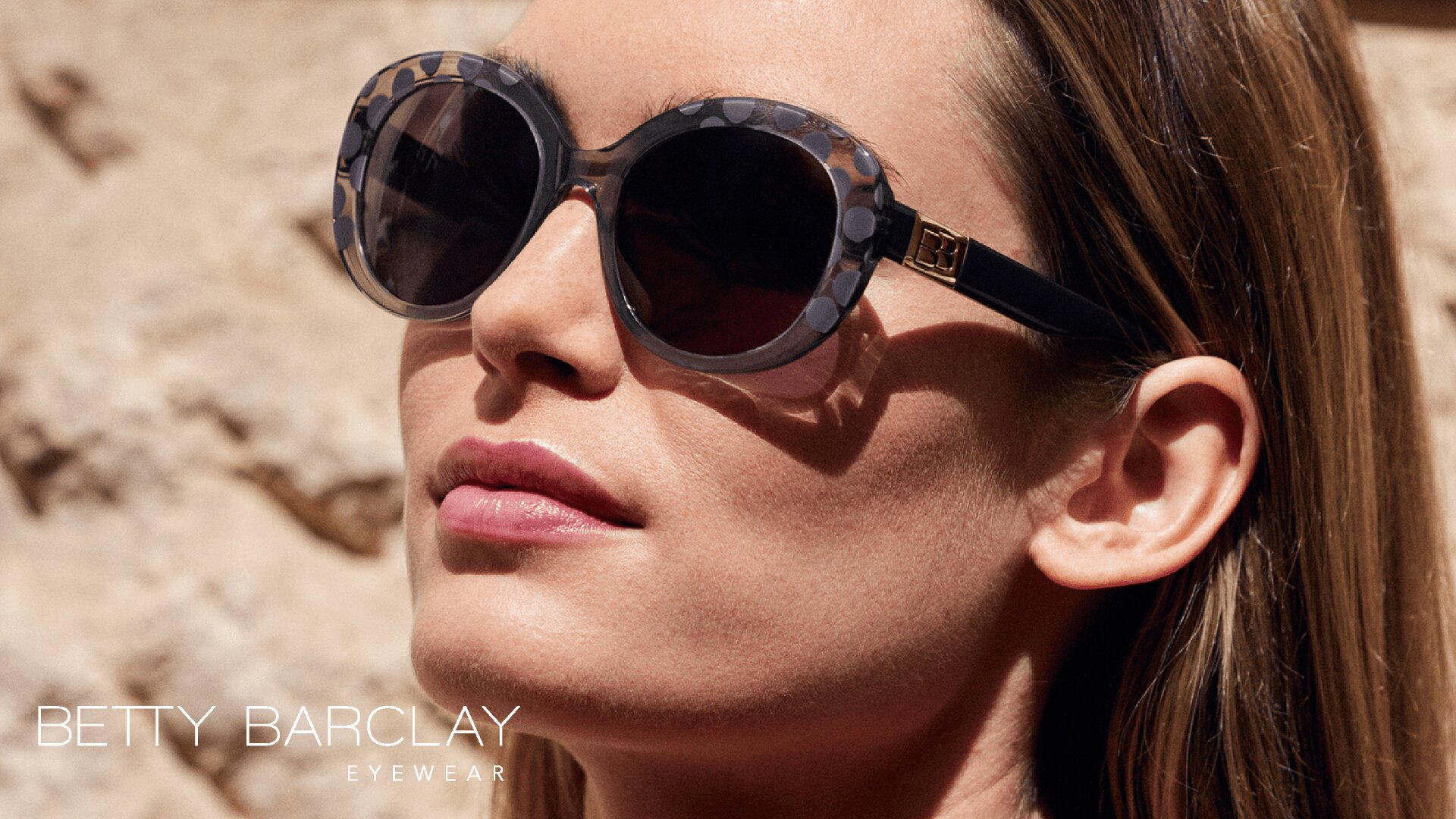 Nahaufnahme einer Frau, die eine große, runde, gemusterte Sonnenbrille mit schwarzen Bügeln trägt. Sie steht im Freien und hat Sonnenlicht im Gesicht. In der unteren Ecke steht BETTY BARCLAY EYEWEAR.