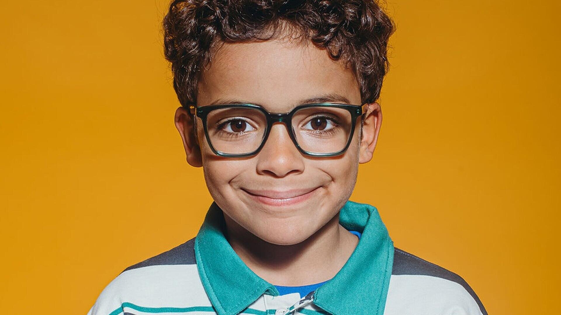 A young boy with curly hair and glasses smiles at the camera against a bright orange background. He is wearing a collared shirt with green, white, and blue stripes.