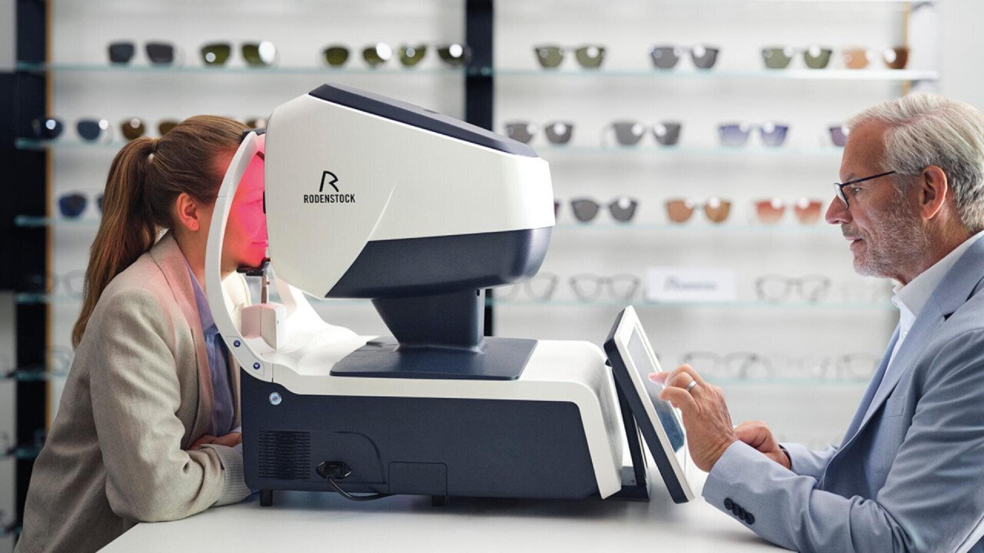 A woman undergoes an eye examination using a modern eye-testing machine, operated by a male optometrist in a clinic with shelves of eyeglasses in the background.