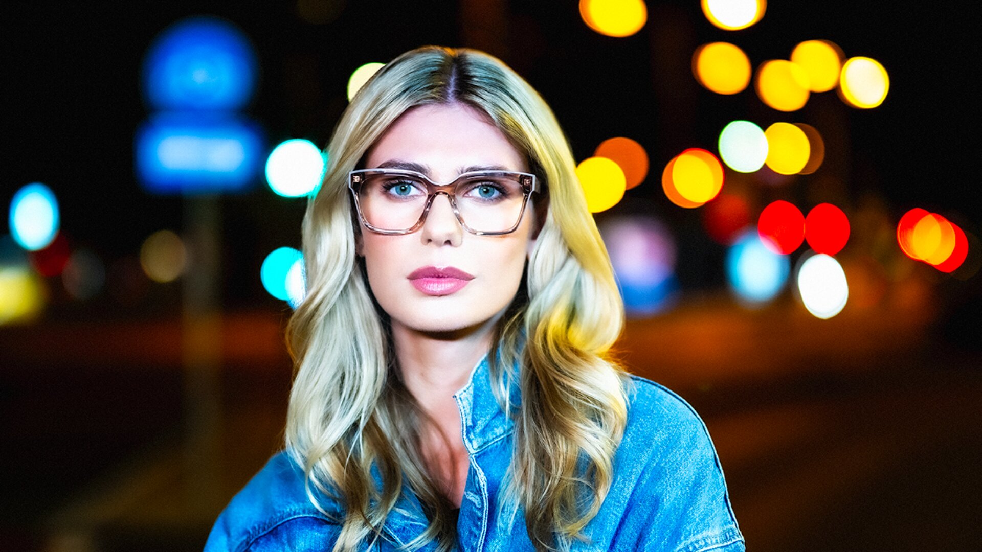 A woman with long blonde hair and glasses wears a denim jacket, standing outdoors at night with colorful, blurred city lights in the background.