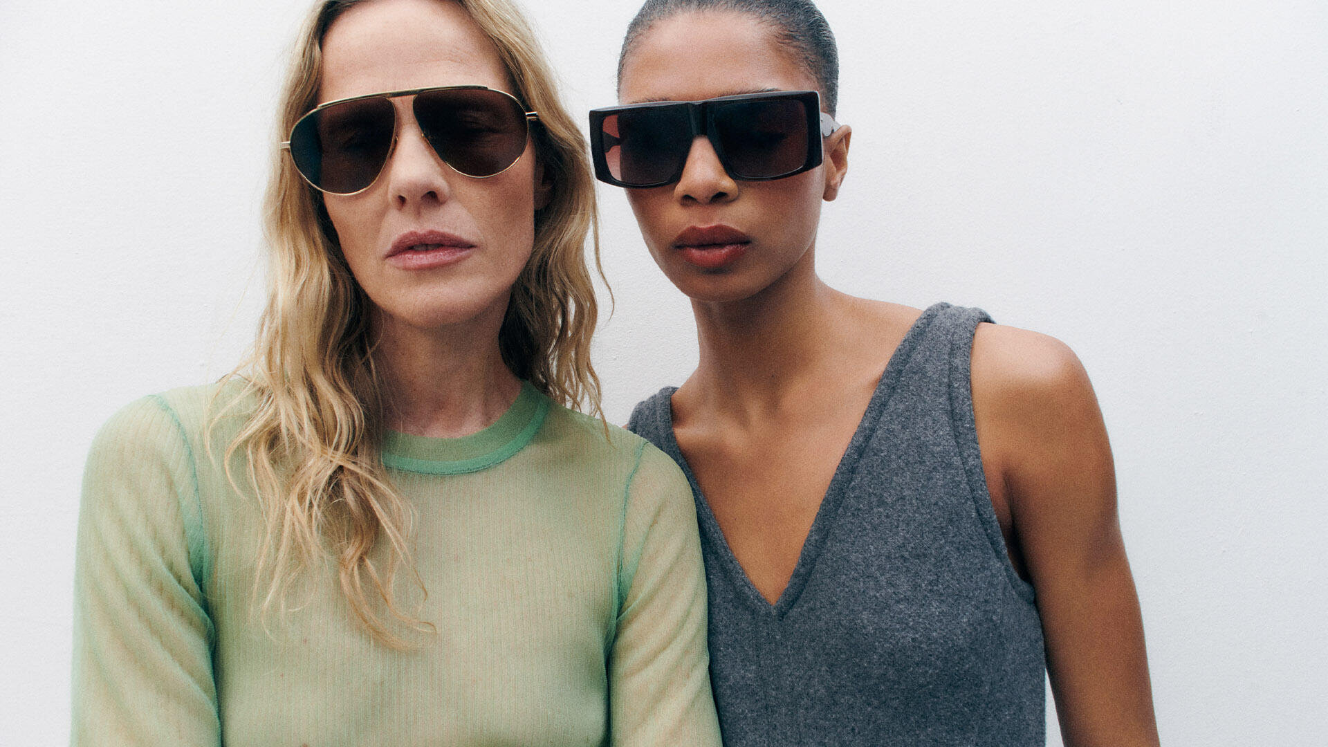 Two women wearing large, dark sunglasses stand side by side against a plain white background. One has long blonde hair and wears a sheer green top; the other has dark skin, slicked-back hair, and wears a sleeveless grey top.