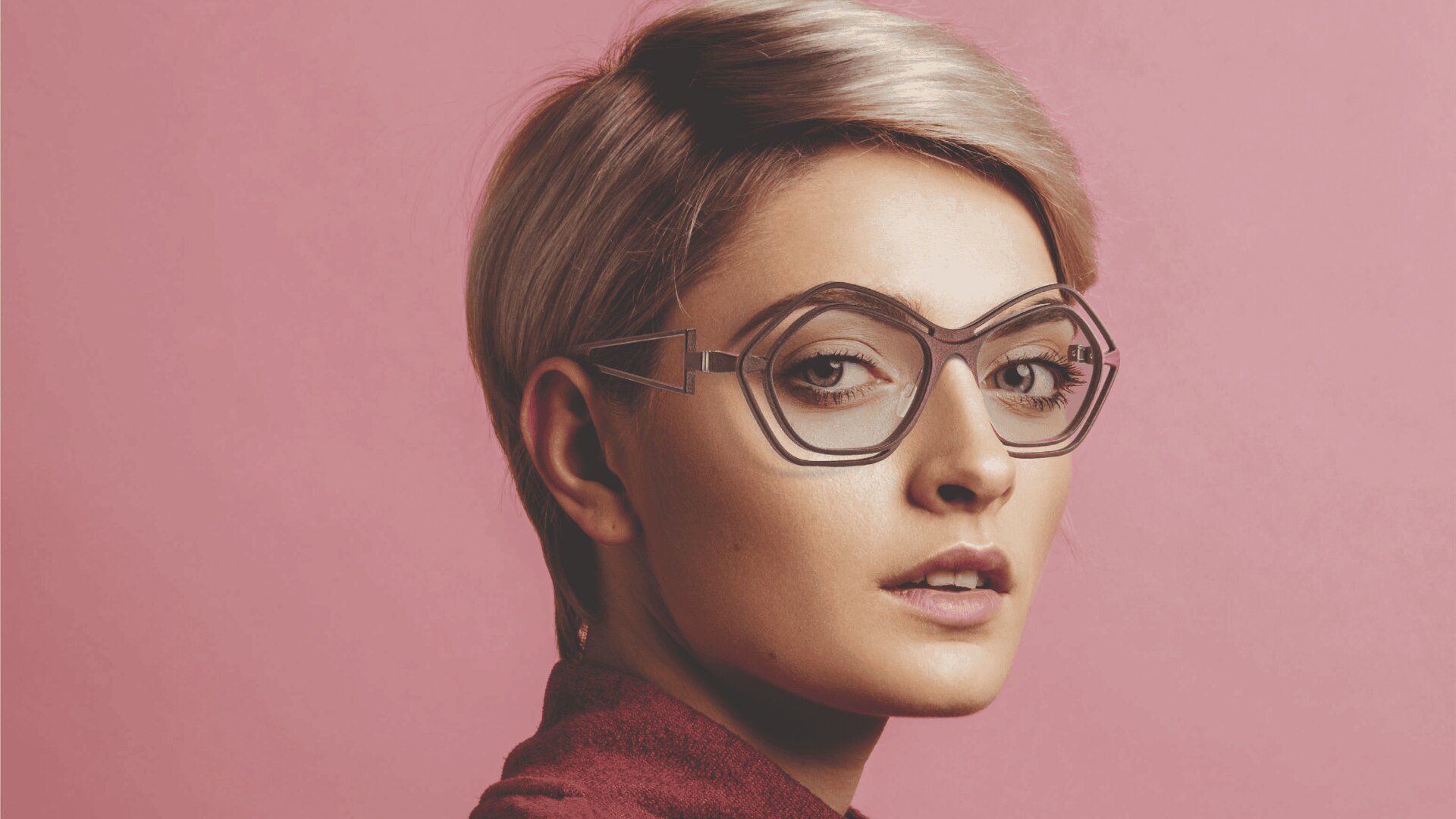 Eine junge Person mit kurzem, hellbraunem Haar blickt über die Schulter, trägt eine übergroße, eigenwillig geformte Brille und ein kastanienbraunes Oberteil vor einem schlichten rosa Hintergrund.