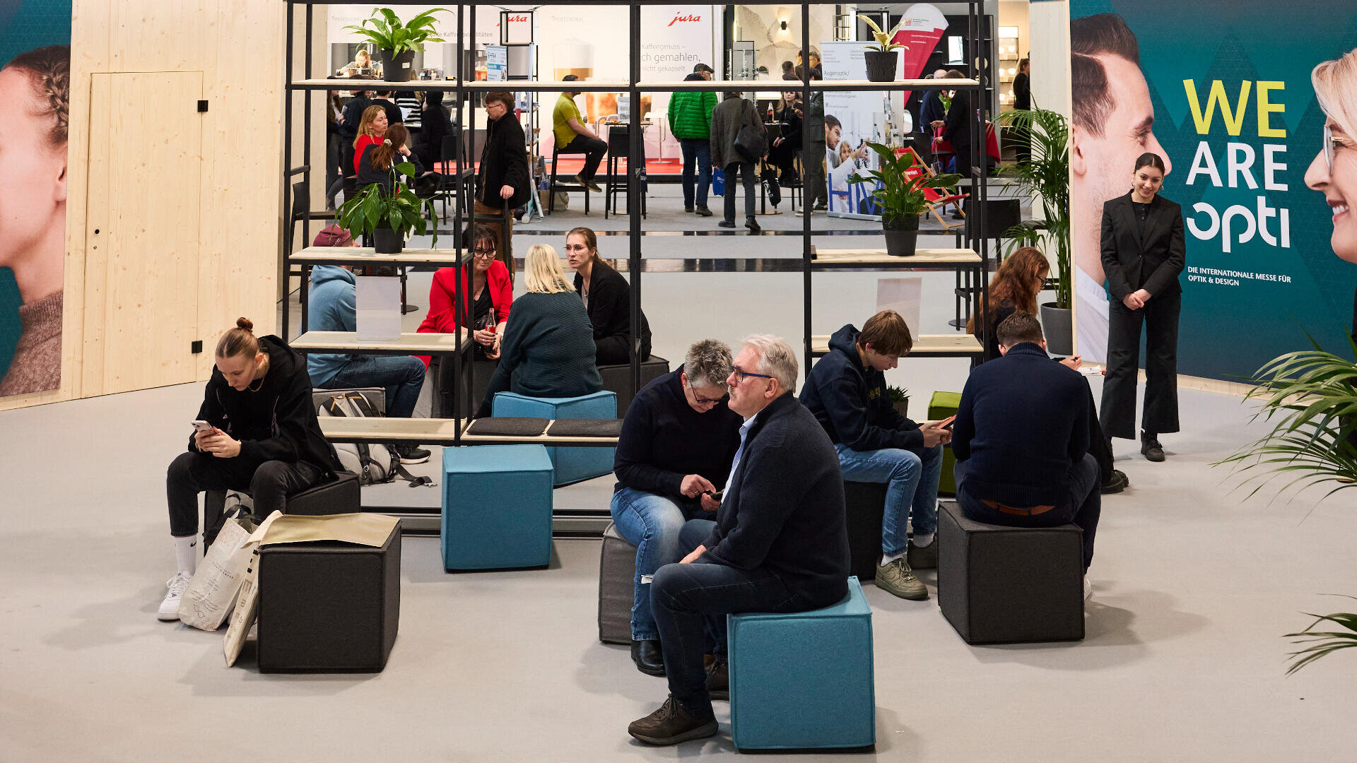 In einem hellen Ausstellungsraum mit Pflanzen und Holzwänden sitzen Menschen auf modernen Sitzwürfeln und unterhalten sich. Im Hintergrund laufen andere umher, auf einem großen Schild steht: „WIR SIND opti“.