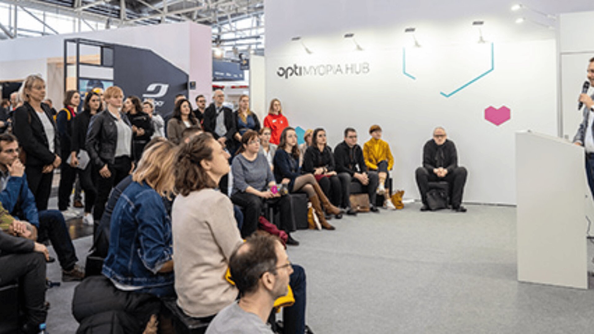 Eine Gruppe von Menschen sitzt und steht aufmerksam da und hört einem Redner bei einer Präsentation in einem modernen Veranstaltungsraum mit OPTIMORAPHUB-Branding an der weißen Wand hinter ihnen zu.