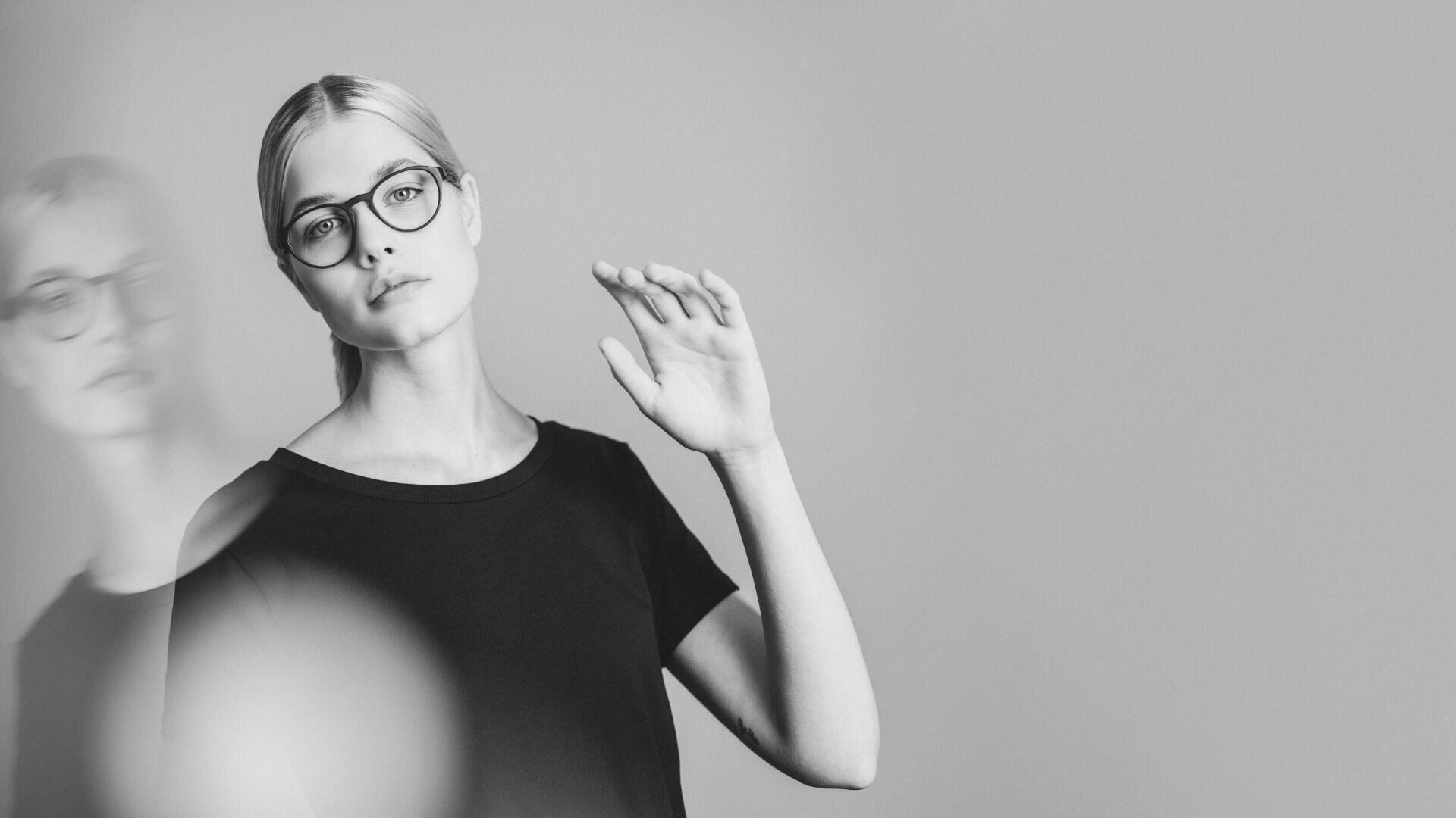 Eine Frau mit hellem Haar, Brille und schwarzem Hemd steht vor einem schlichten Hintergrund. Sie hebt eine Hand, ihr Spiegelbild ist schwach neben ihr zu erkennen. Das Bild ist schwarz-weiß.