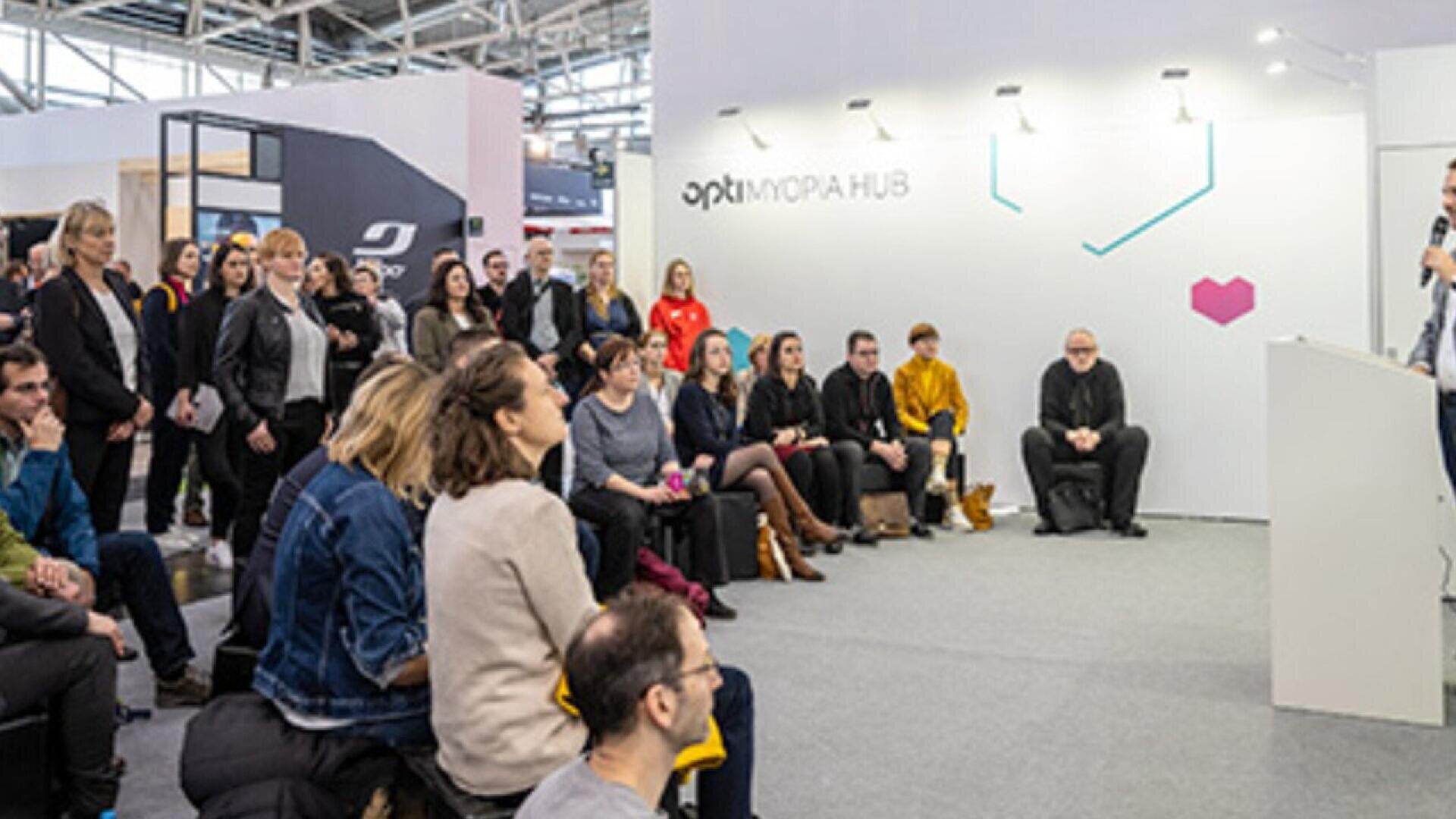 A group of people sit attentively, facing a speaker at a podium in a modern exhibition space labeled OPTI MORIA HUB. Some attendees are standing, and the atmosphere is professional and focused.