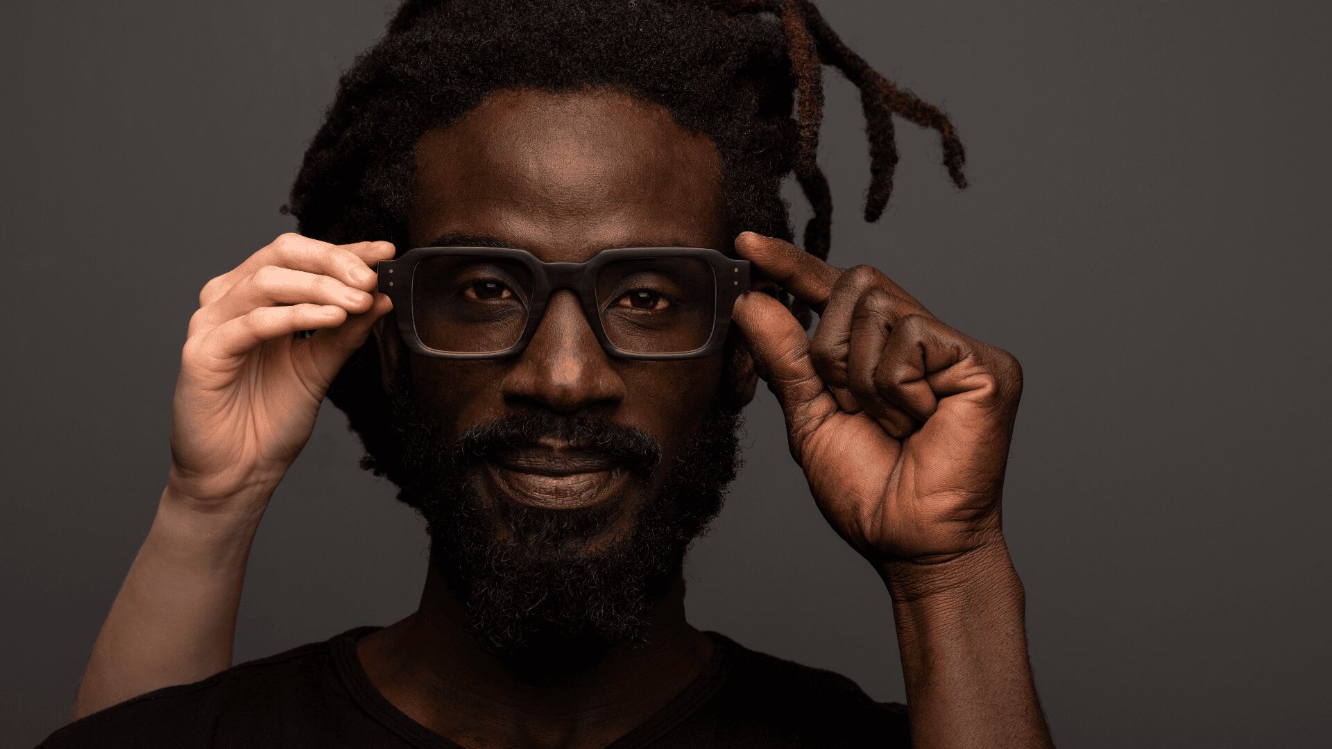 Ein Mann mit dunkler Haut, Bart und Dreadlocks rückt eine große schwarze Brille zurecht, während die Hände einer anderen Person mit hellerer Haut dabei helfen, die Brille vor einem schlichten dunklen Hintergrund auf seinem Gesicht zu positionieren.