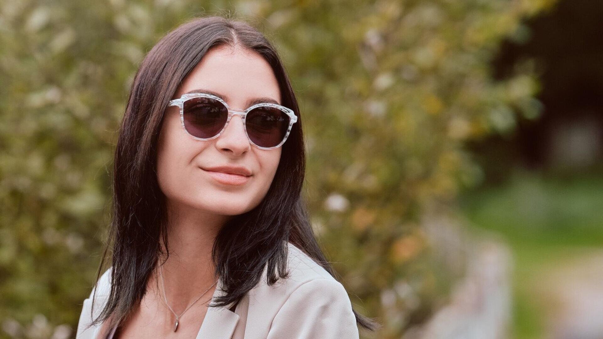 A woman with straight dark hair wearing sunglasses and a light-colored blazer smiles slightly while standing outdoors with a blurred green background.