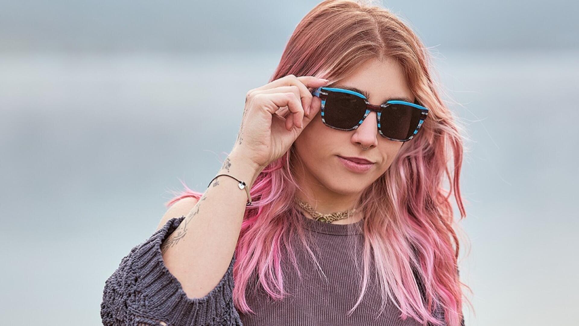 Eine Person mit langen, gewellten rosa Haaren, die eine schwarze Sonnenbrille und einen grauen Pullover trägt, steht im Freien, die Sonnenbrille in einer Hand haltend, mit einem ruhigen, verschwommenen Hintergrund im Hintergrund.