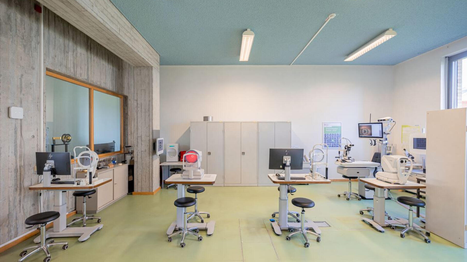 Ein heller, moderner Augenuntersuchungsraum mit mehreren Sehtestgeräten, Hockern, Schränken und einem großen Fenster, das viel Tageslicht hereinlässt. Der Raum hat einen hellgrünen Boden und Betonwände.