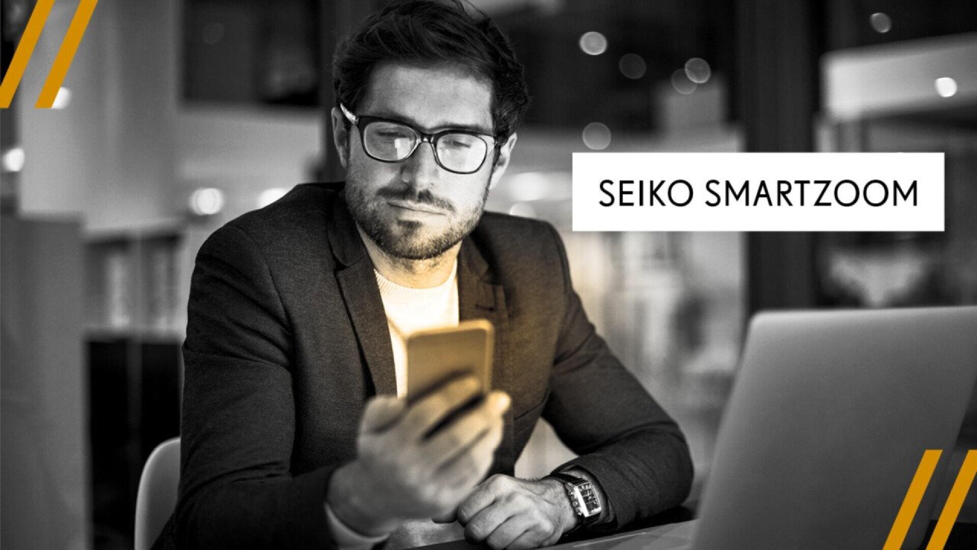 A man in a suit and glasses sits at a desk, looking at his smartphone with a laptop nearby. Text on the image reads SEIKO SMARTZOOM. The photo has a professional and modern feel.