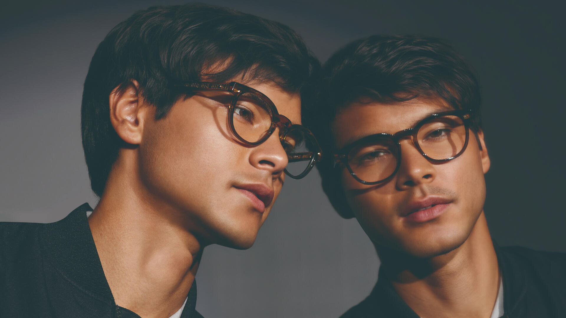 Ein junger Mann mit Brille lehnt seinen Kopf an einen Spiegel, wodurch sein Gesicht reflektiert wird. Er hat kurzes dunkles Haar und blickt nachdenklich zur Seite. Der Hintergrund ist sanft und neutral beleuchtet.