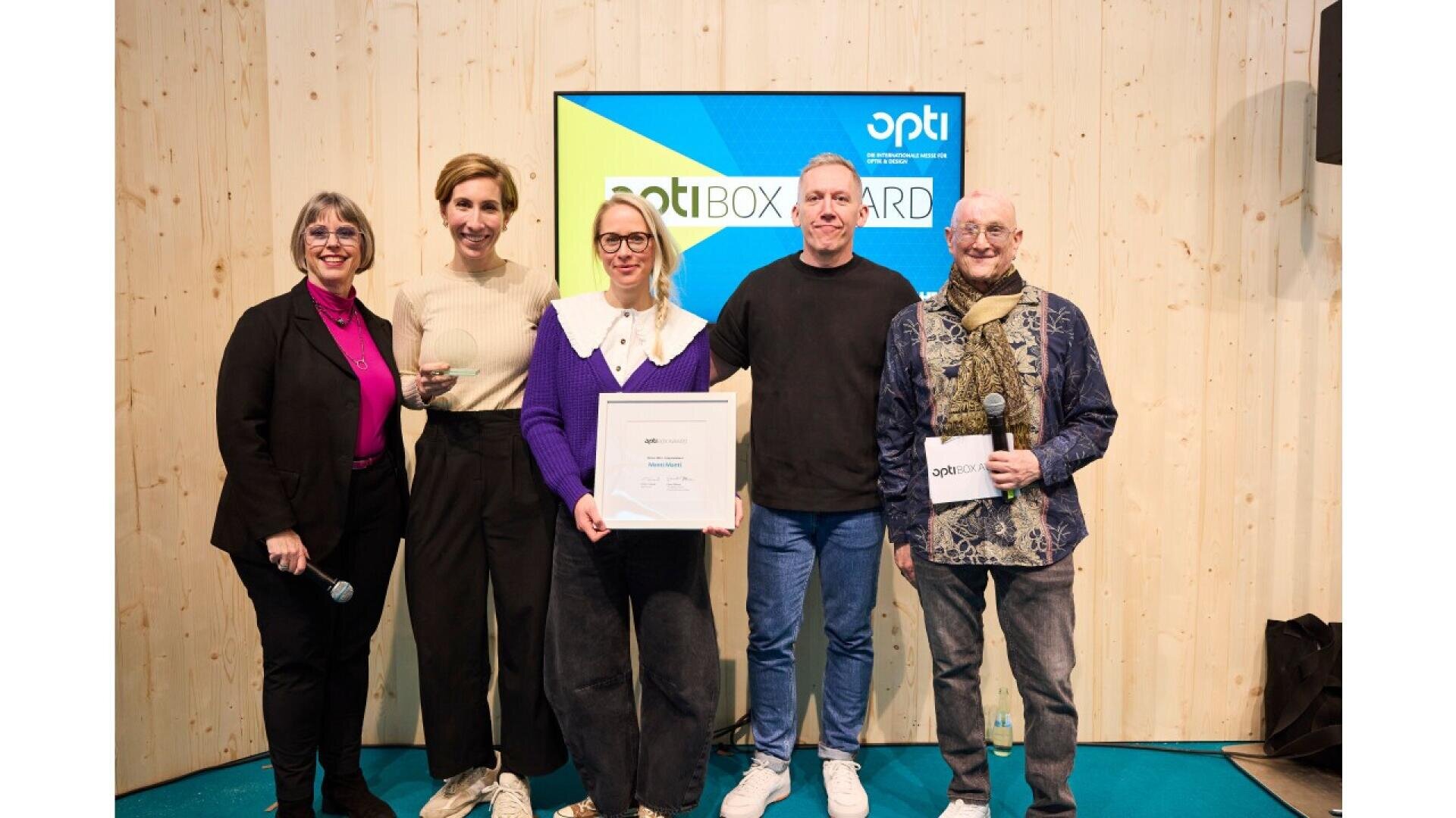Five people stand in front of a wooden wall and a screen displaying opti BOX AWARD. The person in the center holds a framed certificate, and two others are holding microphones. All are smiling.