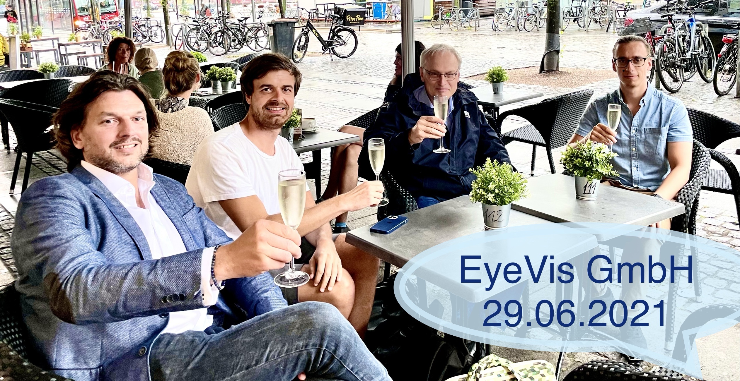 Four men sit at an outdoor café table, smiling and toasting with a glass of champagne. The scene is relaxed and casual. A blue text overlays EyeVis GmbH 29.06.2021. Bicycles and people can be seen in the background.