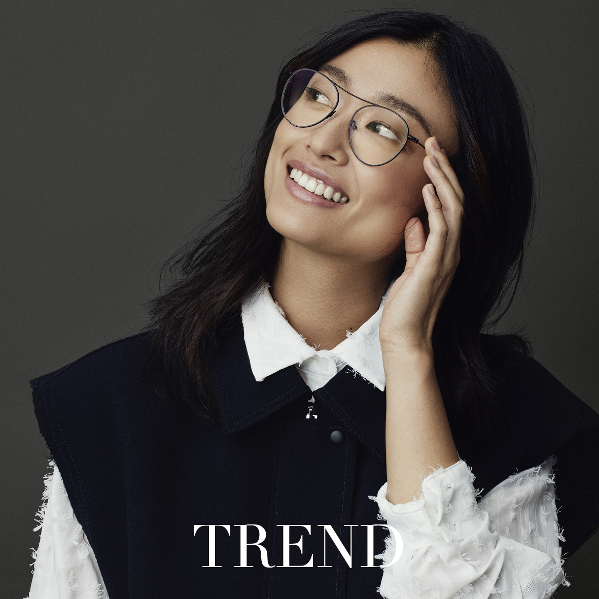 Eine Frau mit langen dunklen Haaren und Brille lächelt und berührt ihr Gesicht. Sie trägt ein weißes Hemd mit Kragen und eine schwarze Weste. Am unteren Rand des Bildes ist das Wort TREND zu lesen. Der Hintergrund ist dunkelgrau.