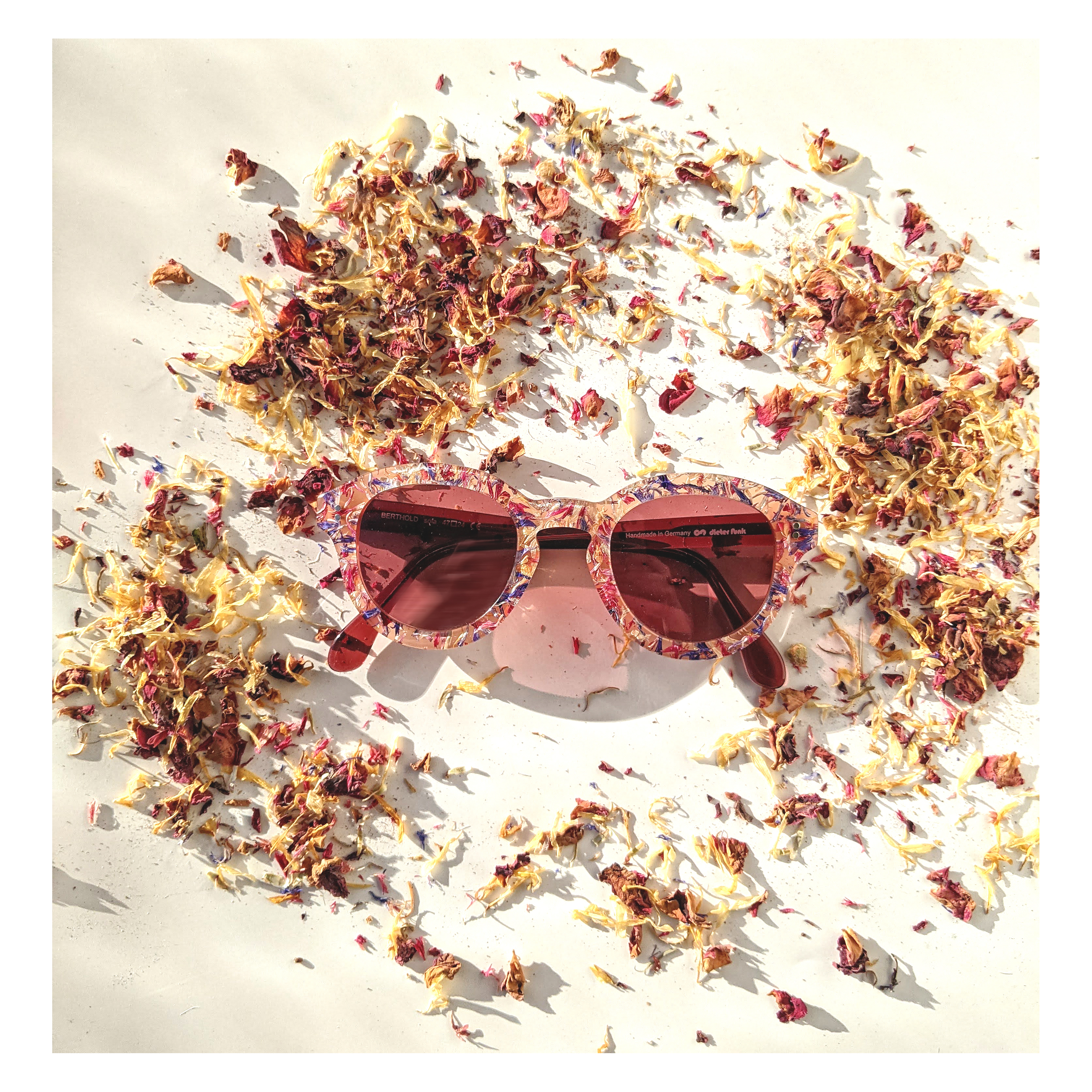 Eine rosa Sonnenbrille mit floralem Muster liegt auf einer weißen Oberfläche, umgeben von verstreuten getrockneten Blütenblättern in Rot-, Gelb- und Beigetönen, wodurch eine lebendige, sommerliche Komposition entsteht.