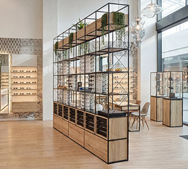 Modern eyewear store with wooden floors, open eyewear shelves, hanging plants and large windows that let in natural light. The space has a clean, minimalist and organized design.