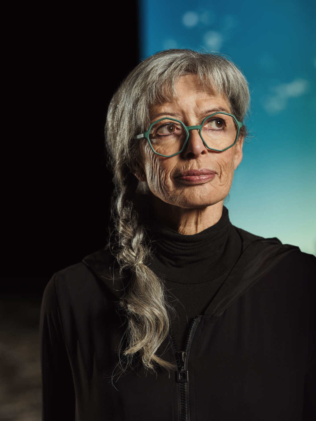 Eine ältere Frau mit langen grauen Haaren, die zu einem Zopf geflochten sind, trägt eine blaugrüne Brille und ein schwarzes Oberteil und blickt nachdenklich zur Seite. Der Hintergrund ist halb dunkel und halb blau mit einem weichen, unscharfen Effekt.