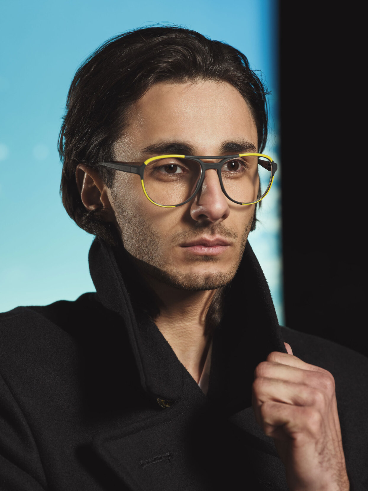 Ein junger Mann mit mittellangem, dunklem Haar trägt eine große, modische Brille mit gelben Rändern und einen schwarzen Mantel, dessen Kragen hochgeschlagen ist. Der Hintergrund weist einen Farbverlauf von Blau und Schwarz auf.