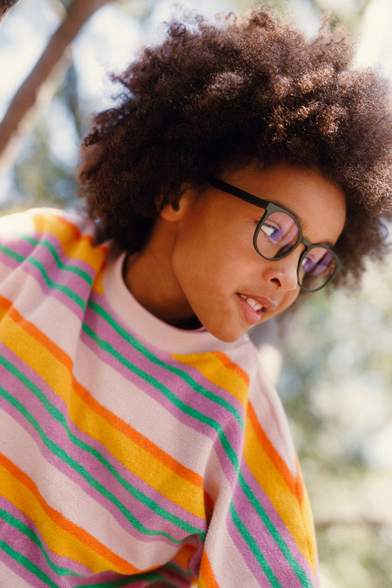 Ein Kind mit lockigem Haar und Brille trägt einen bunten, gestreiften Pullover und blickt mit einem sanften Lächeln nach unten, im Freien mit unscharfem Grün im Hintergrund.
