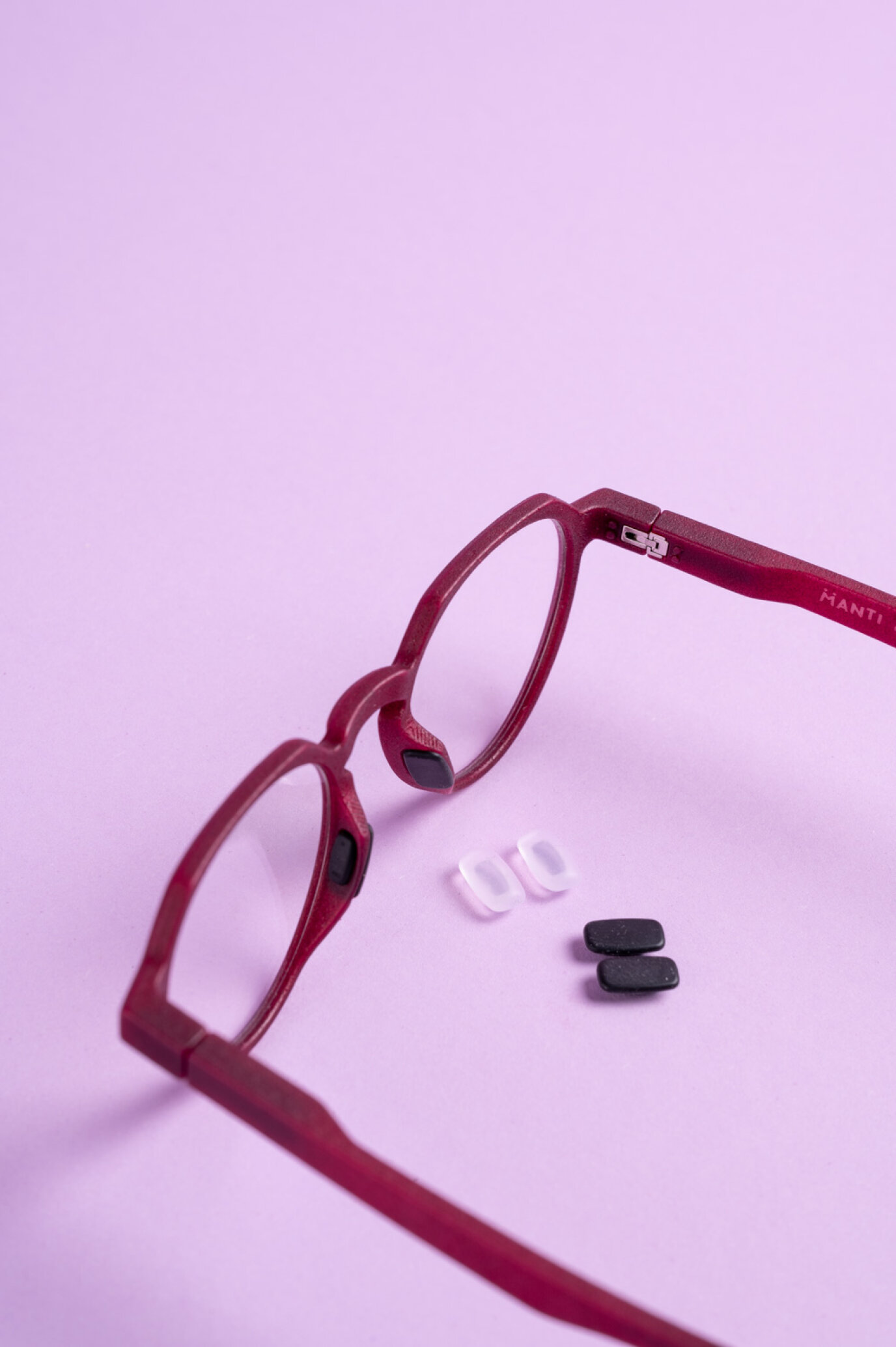 Rote Brille mit abnehmbaren schwarzen und weißen Nasenpads auf einer hellvioletten Oberfläche.