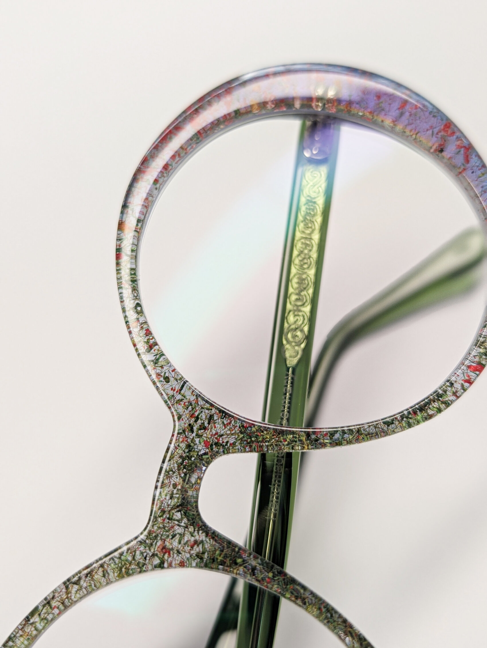 Nahaufnahme einer runden Brille mit einem gesprenkelten, mehrfarbigen Rahmen und einem verschlungenen goldenen Muster auf der Innenseite des Bügels vor einem schlichten hellen Hintergrund.