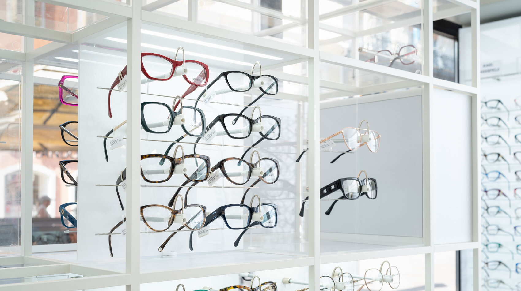 Eine Auslage mit verschiedenen Brillen mit unterschiedlichen Fassungen und Farben, ordentlich auf weißen Regalen in einem Optikergeschäft angeordnet.