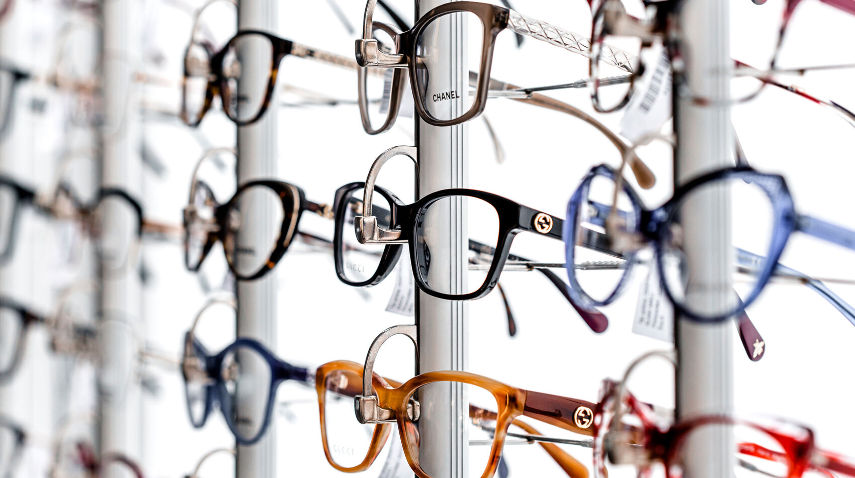 Ein Regal mit verschiedenen Brillen in unterschiedlichen Formen, Größen und Farben, darunter Marken wie Chanel und Gucci, die alle in Reihen vor einem weißen Hintergrund angeordnet sind.