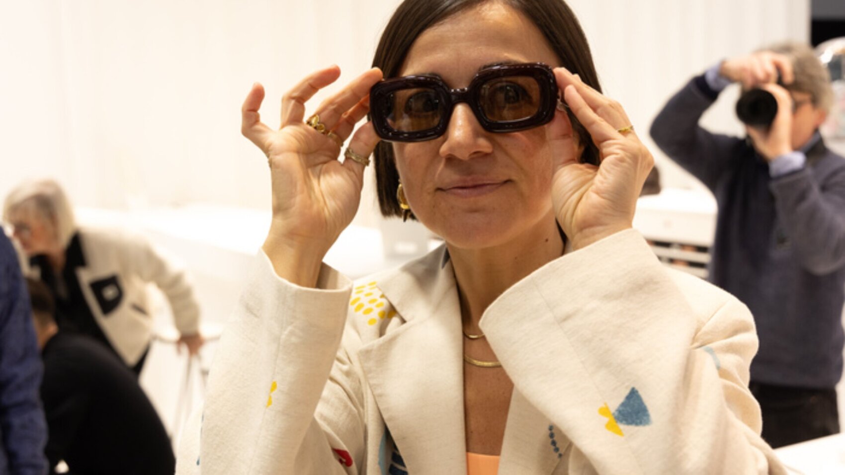 A woman wearing a light-patterned blazer smiles while holding oversized sunglasses to her face. Other people are visible in the blurred background, including someone taking a photo.