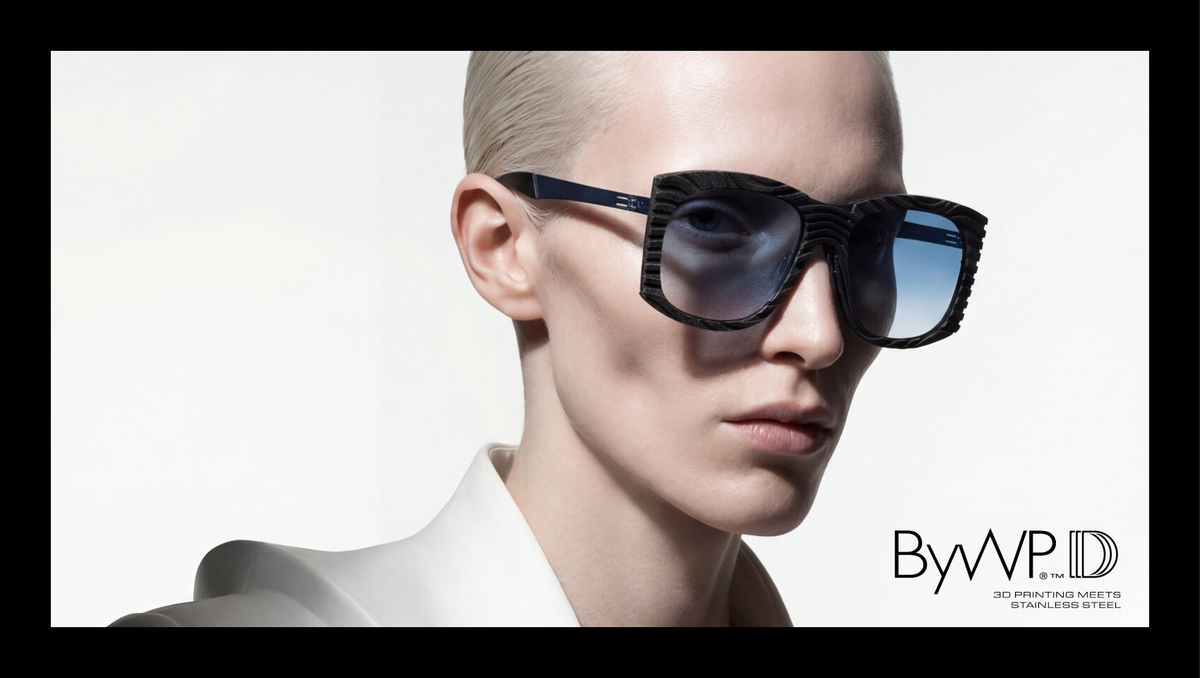 A person with short, slicked-back blond hair wears large, geometric blue sunglasses and a white collared shirt against a light background. At the bottom right it says: ByWP: 3D printing meets stainless steel.