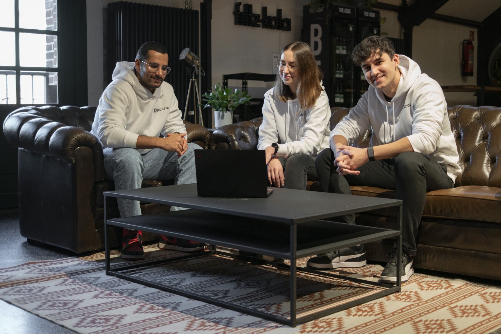 Drei junge Erwachsene in weißen Kapuzenpullovern sitzen zusammen auf braunen Ledersofas um einen schwarzen Couchtisch mit einem Laptop, lächeln und unterhalten sich in einem gemütlichen, modernen Raum mit Pflanzen und warmer Beleuchtung.