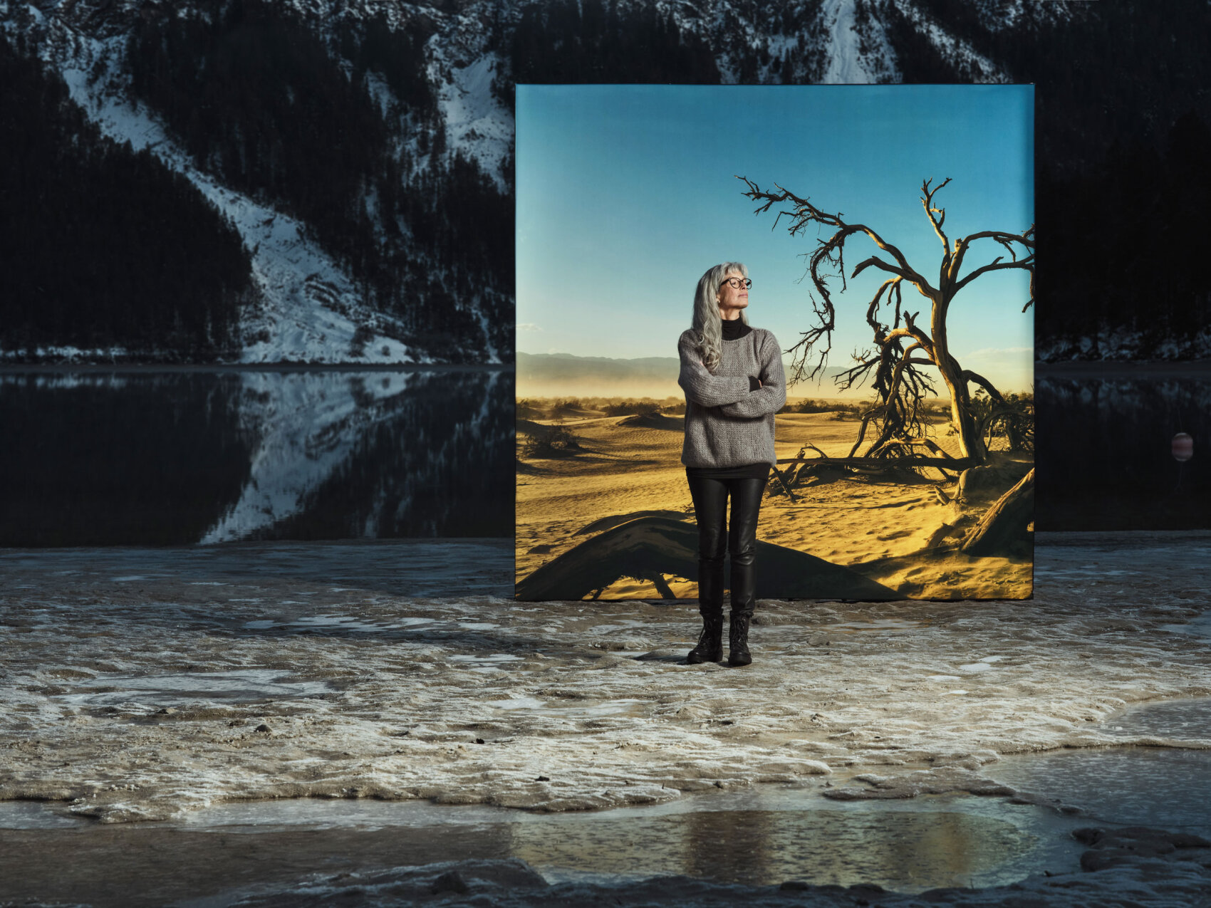 Eine Frau in grauem Pullover und schwarzer Hose steht auf einer schneebedeckten Fläche vor dem Hintergrund einer trockenen, sonnenbeschienenen Landschaft mit einem kahlen Baum, der mit verschneiten Bergen und einem See im realen Hintergrund kontrastiert.